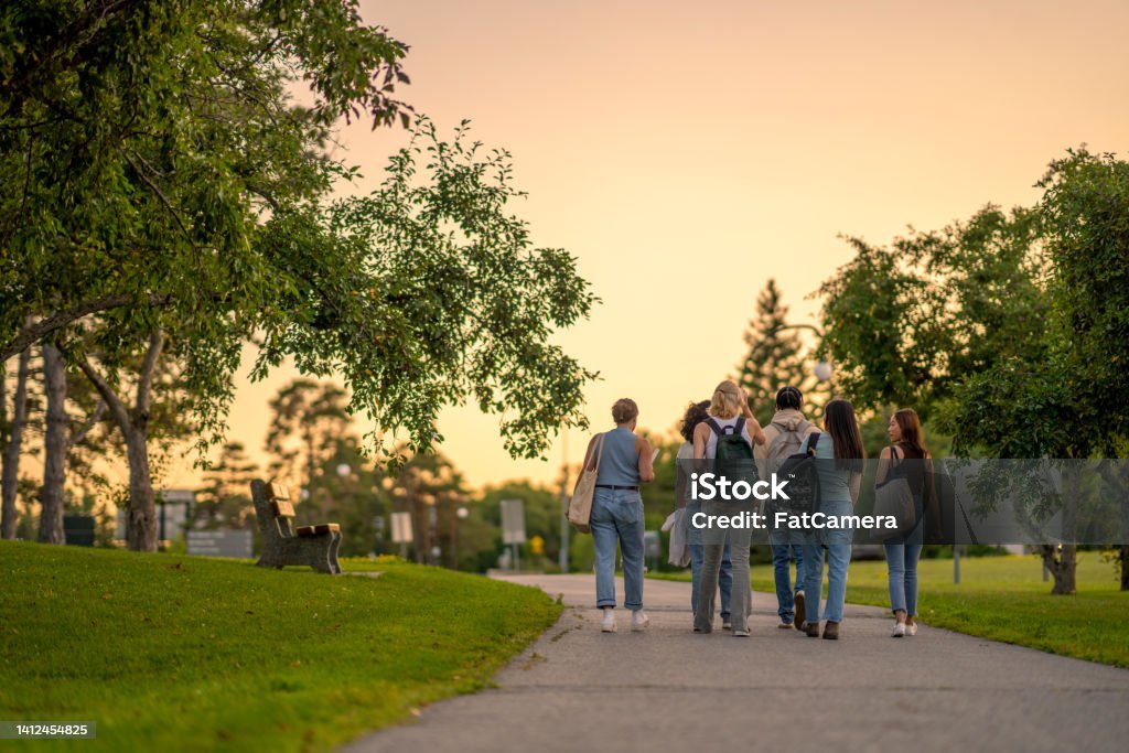 A group of University students are seen from behind walking outside on campus as they make their way to class.  They are dressed casually and have backpacks on as they talk together and make their way to school for the day.